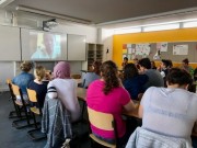Das Bild zeigt eine Gruppe von Menschen, die in einem Lernraum zusammensitzen und auf ein Smartboard blicken, auf dem ein Skypeinterviewpartner eingeblendet ist.