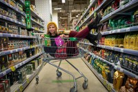Das Bild zeigt eine junge Frau, die beladen mit Einkäufen in einem Einkaufswagen zwischen zwei Supermarktregalen sitzt und in die Kamera lächelt.