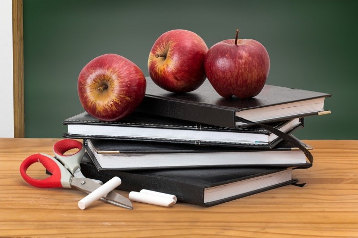 Das Bild zeigt gestapelte Bücher und Hefte, eine Schere, Kreide und drei Äpfel.