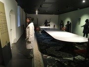 Das Bild zeigt einen Museumsraum mit Ritterrüstungen und einer großen Festtafel. Eine Frau steht am Rand und betrachtet eine Infotafel.