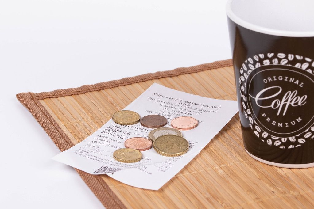 Das Bild zeigt eine Tasse Kaffee mit Rechnung und Kleingeld.